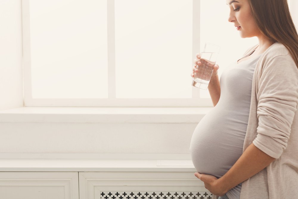 Pregnant woman drinking water, standing at window. Young expectant lady have rest at home with glass, copy space. Pregnancy, healthcare, thirst concept